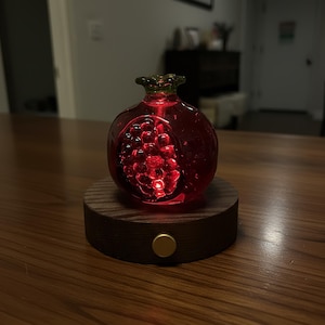 May include: A decorative, red glass pomegranate with a green stem, illuminated from within. The pomegranate sits on a round, dark wood base with a gold-colored button. The interior of the pomegranate is filled with red, glowing orbs.