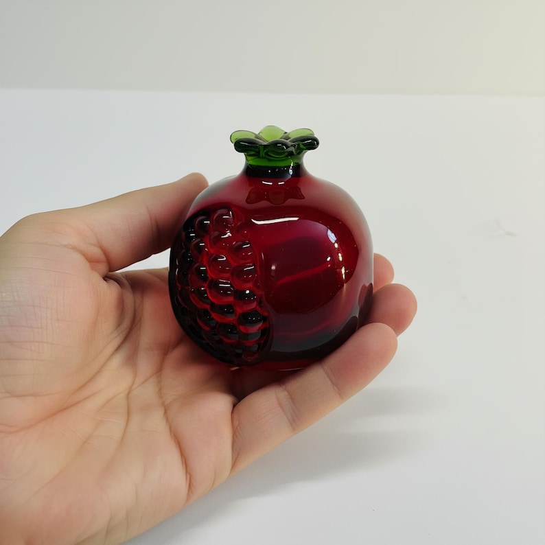 May include: A small, decorative glass pomegranate in a deep red hue, with a green stem and a textured, seed-like design on one side. The fruit is held in a hand, showcasing its size and detail.