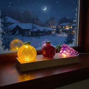 Puede incluir: Luces decorativas de frutas de cristal, incluyendo una naranja amarilla, una granada roja y un racimo de uvas moradas, sobre una repisa de madera. Las luces iluminan una escena de noche de invierno nevada.