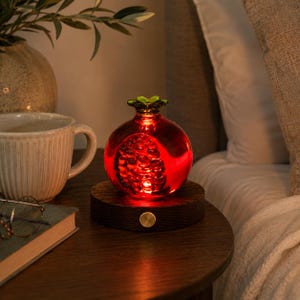May include: A red, pomegranate-shaped decorative lamp with a green top, illuminated from within. It sits on a dark wooden base with a gold-colored button. The lamp is on a wooden nightstand next to a coffee mug and a book.