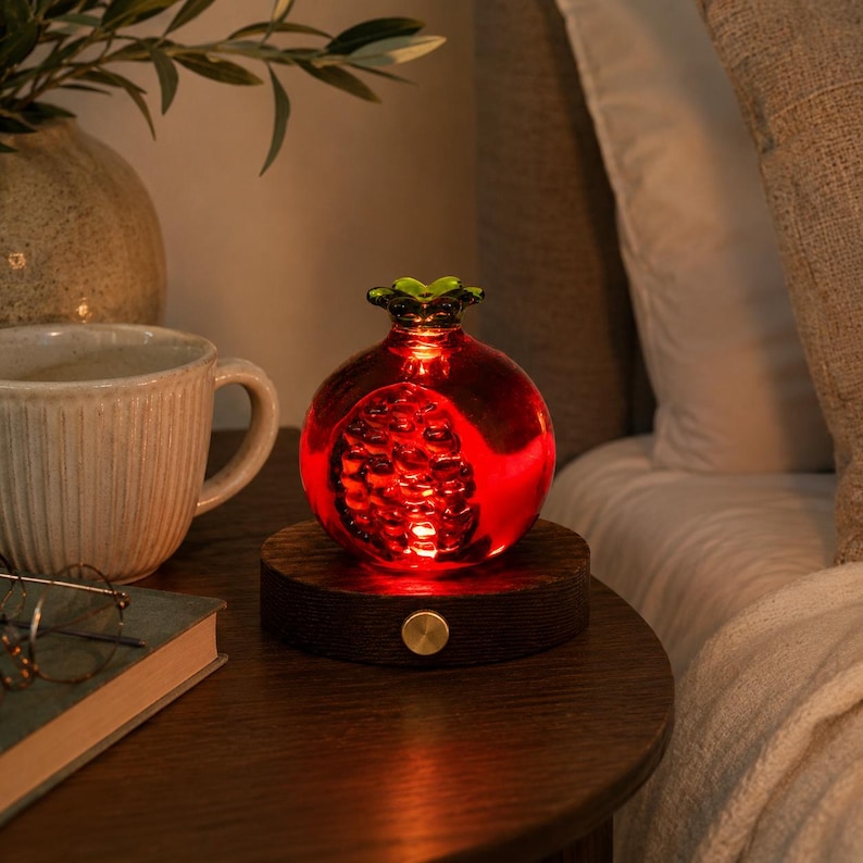 May include: A red, pomegranate-shaped decorative lamp with a green top, illuminated from within. It sits on a dark wooden base with a gold-colored button. The lamp is on a wooden nightstand next to a coffee mug and a book.