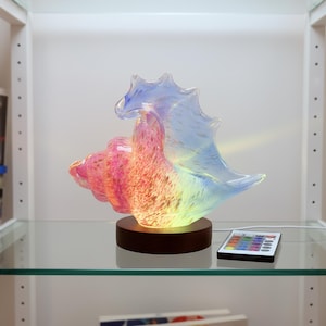 Könnte beinhalten: Eine farbenfrohe Glas-Muscheln-Skulptur mit Holzsockel. Die Skulptur wird mit einem Regenbogen von Farben beleuchtet, und eine Fernbedienung liegt daneben. Die Muschel ist ein dekoratives Element.