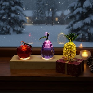 Puede incluir: Una exhibición festiva con esculturas de frutas de vidrio iluminadas: una manzana roja, una pera morada y una piña amarilla. Una pequeña caja de regalo roja con una cinta dorada también está presente. La escena se desarrolla contra un fondo invernal nevado.