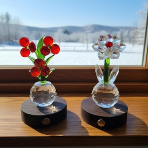 Puede incluir: Dos esculturas decorativas de flores de cristal. Una presenta bayas de cristal rojo y hojas verdes, la otra una flor transparente con un centro rojo. Ambas se asientan sobre bases de madera oscura con un acento dorado.