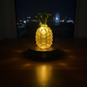 May include: A glowing pineapple-shaped lamp with a textured surface and green leafy top. The lamp sits on a dark wooden base with a small button. The background shows a blurred cityscape at night.