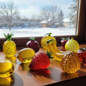 Può includere: Una collezione di sculture di frutta in vetro colorato, tra cui un ananas, un limone, una fragola, banane e mele, è disposta su una superficie di legno. Le sculture di frutta sono nelle tonalità del giallo, rosso e verde, con una scena esterna innevata visibile attraverso una finestra sullo sfondo.
