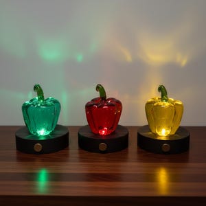 May include: Three glass bell pepper-shaped decorative lights in green, red, and yellow. Each pepper sits on a dark wooden base with a small gold-colored button. The lights illuminate the peppers from within, creating a colorful display.