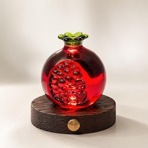 Pode incluir: Uma romã decorativa de vidro vermelho vibrante, com um interior detalhado. Assenta sobre uma base de madeira escura com um detalhe em latão. O caule verde da romã adiciona um toque de contraste.