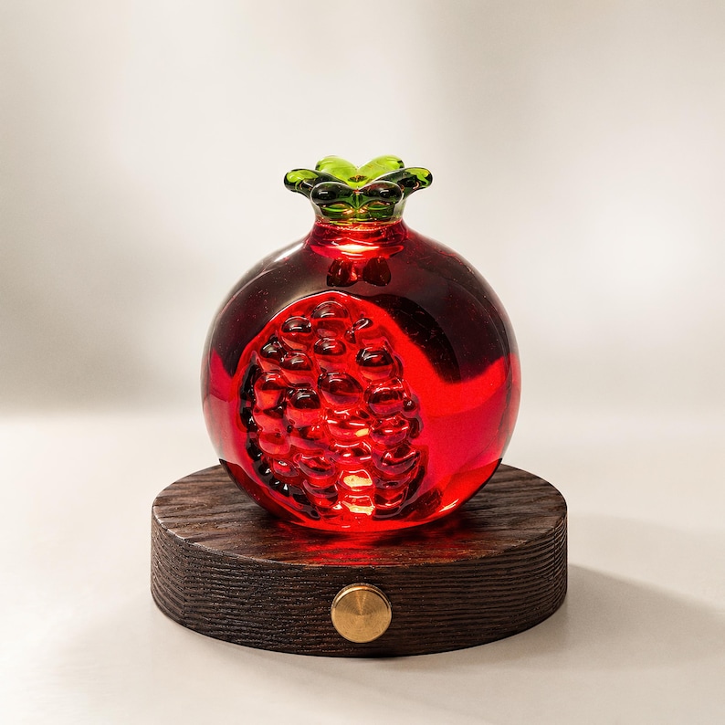 May include: A decorative glass pomegranate with a vibrant red hue, showcasing a detailed interior. The fruit sits atop a dark wooden base with a brass accent. The pomegranate's green stem adds a contrasting touch.