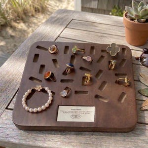 Puede incluir: Exhibición de anillos y una pulsera de perlas en una bandeja de madera. Los anillos presentan diferentes piedras preciosas y diseños, incluido un anillo en forma de trébol. La bandeja tiene formas recortadas y una pequeña etiqueta con texto.