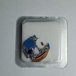 May include: A watch movement component, featuring a blue circuit board with gold and red accents. The component is encased in a clear plastic package, set against a white background. This item is likely a replacement part for watch repair.