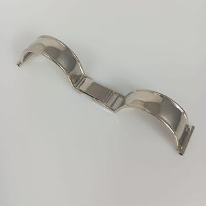 Op de afbeelding: Een gepolijste zilverkleurige metalen horlogeband met een gebogen, halfstijf ontwerp en een centraal vouwsluitingsmechanisme. De band bestaat uit twee symmetrische, gladde en reflecterende gebogen segmenten die in het midden verbonden zijn.