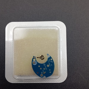 May include: A small, blue circuit board with gold-coloured components is encased in a clear plastic package. The board has a semi-circular shape with several circular components and a small, gold-coloured metal piece attached.