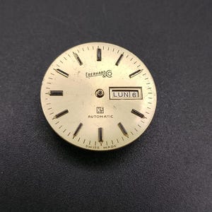 May include: A gold-coloured watch face with black hour markers and hands. The face displays the brand name "Eberhard & Co" and the day of the week, "LUN 6". The words "Automatic" and "Swiss Made" are also visible.