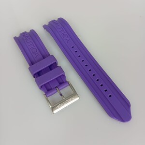 Könnte beinhalten: Ein violettes Silikon-Uhrenarmband bestehend aus zwei Teilen. Das Armband zeigt das Wort NAUTICA, das auf dem Band eingeprägt und auf der silberfarbenen Metallschnalle eingraviert ist. Das Band enthält zwei verschiebbare Schlaufen und Einstelllöcher.
