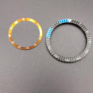 May include: Two circular watch bezel inserts. The smaller insert is orange with white numerals 13 through 23. The larger insert is black with white numerals 5 through 40, featuring a blue section from 5 to 10 and a white triangle marker at the top.