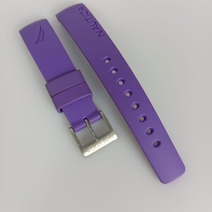 Könnte beinhalten: Ein violettes Silikon-Uhrenarmband mit dem eingeprägten Nautica-Logo auf dem Band und einer Gravur auf der silberfarbenen Metallschnalle. Das Armband enthält eine passende violette Silikonschlaufe und eine Reihe runder Einstelllöcher.
