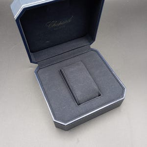 Peut inclure: Boîte de montre bleu marine ouverte, avec un intérieur bleu foncé et un support pour montre. Le nom de la marque "Chopard" est visible à l'intérieur du couvercle. Bordure argentée.