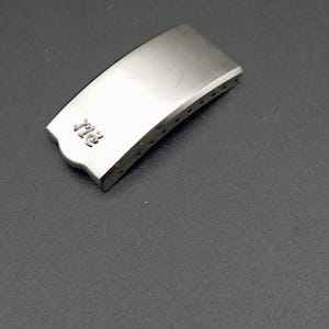 Può includere: Una chiusura per orologio in metallo color argento con le lettere "RC" incise sulla parte superiore. La chiusura ha una forma rettangolare curva con diversi piccoli fori lungo un bordo. La superficie è liscia e riflettente.
