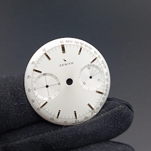 May include: A white watch face with black markings and the brand name "ZENITH". It features two sub-dials, a central hole, and various numerical indicators. The face is held by a gloved hand.