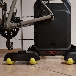 Könnte beinhalten: Ein schwarzer Heimtrainer mit einem silbernen Fahrrad. Der Trainer hat eine schwarze Basis mit gelben Rädern. Kette und Zahnräder des Fahrrads sind sichtbar. Für Indoor-Radtraining konzipiert.