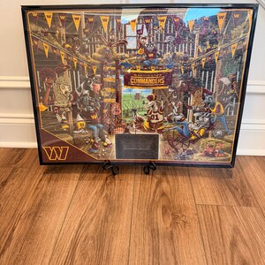 May include: Framed artwork featuring a detailed illustration of Washington Commanders team characters in a stadium setting. The artwork has a black frame and is displayed on a wooden floor. The team's logo and name are prominently displayed.