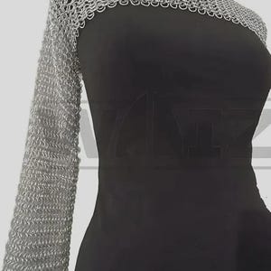 Puede incluir: Una hombrera de cota de malla plateada con una manga larga, exhibida en un maniquí negro. La cota de malla está hecha de pequeños anillos metálicos entrelazados. El maniquí es negro y tiene una forma simple y elegante.