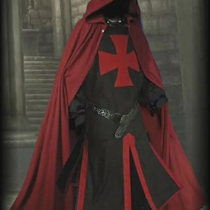 Pode incluir: Um traje medieval com uma longa capa vermelha com capuz e uma túnica preta com um emblema de cruz vermelha. A túnica tem acabamentos vermelhos e é usada com um cinto decorativo. O traje está em um fundo de pedra.