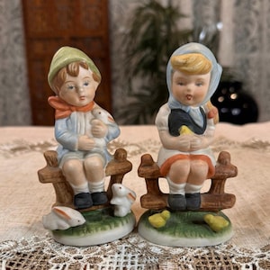 May include: Two ceramic figurines of children sitting on a wooden fence. One holds a duck, the other a chick. Both are wearing vintage clothing. A white rabbit and two yellow chicks are at the base of the figurines.