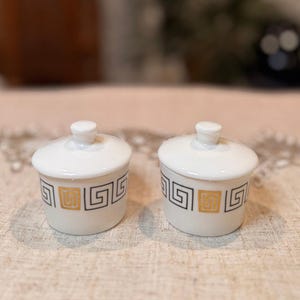 May include: Two small, white ceramic lidded jars. Each jar features a black and gold geometric pattern around the base. The lids have a small, rounded knob. The jars are set on a neutral-toned surface.