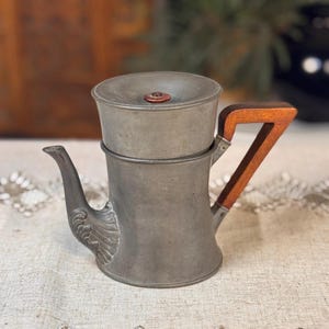 May include: Antique pewter coffee pot with a wooden handle. The pot features a spout with an embossed design and a lid with a small, round, reddish-brown knob. The handle is a dark wood with a light wood accent.