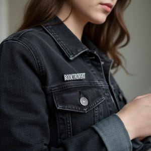 Könnte beinhalten: Schwarze Jeansjacke mit einer silbernen Anstecknadel mit der Aufschrift "BOOKTROVERT" in einer serifenlosen Schriftart. Die Anstecknadel ist rechteckig und hat einen dunklen Hintergrund. Die Jacke hat eine geknöpfte Tasche und einen Knopfverschluss.