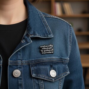 Könnte beinhalten: Eine blaue Jeansjacke mit einem schwarzen T-Shirt darunter. Eine schwarze Anstecknadel mit dem Text "emotionally attached to fictional characters" ist auf die Jacke gesteckt. Die Jacke hat silberne Knöpfe und ein klassisches Design.