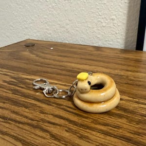 May include: A coiled, tan-coloured snake keyring with a small yellow hat. The snake has black eyes and a white belly. The keyring is made of a shiny material and has a silver clasp.