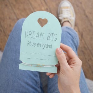 Puede incluir: Una tarjeta azul claro con un recorte en forma de corazón y las palabras "DREAM BIG" y "Rêve en grand" impresas. Una pulsera blanca y dorada está unida a la tarjeta. La tarjeta es sostenida por una persona que lleva vaqueros azules.