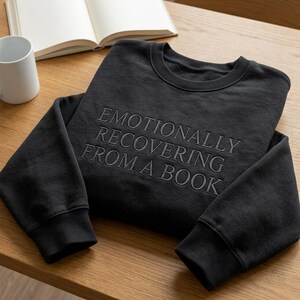 May include: A black long-sleeve crewneck sweatshirt featuring the embroidered text EMOTIONALLY RECOVERING FROM A BOOK in a matching black thread. The garment has a ribbed collar, cuffs, and hem, showcasing a clean and simple design.