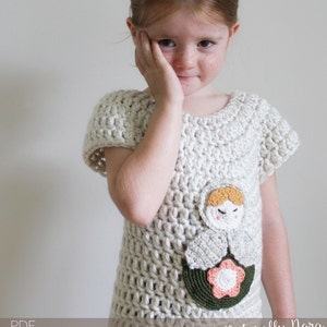 Könnte beinhalten: Ein weißes Häkeltop mit kurzen Ärmeln und einer gehäkelten Puppenapplikation. Die Puppe hat eine rosa Blume und grüne Blätter. Der Text "Naturally Nora Crochet" ist unten im Bild sichtbar.