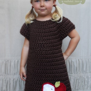 May include: A brown crochet dress with short sleeves and an apple applique. The dress is worn by a young girl with blonde hair and pigtails. The text "PDF Crochet Pattern" is written in a green box at the top of the image. The text "Not a finished crocheted item." is written below the box.