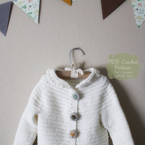 May include: White crocheted cardigan with a hood and four buttons featuring a tree design. The cardigan is hanging on a wooden hanger against a light beige background. The text "PDF Crochet Pattern" is written in green on a white background.