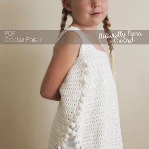 May include: A white crochet top with a scalloped hem and a crocheted flower detail. The top is sleeveless and has a scoop neck. The model is wearing the top with blue denim shorts. The text "Naturally Nora Crochet" is visible in the image.