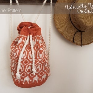 Puede incluir: Una mochila de crochet naranja y blanca con cordones blancos que cuelgan de la parte superior. La mochila está colgada de un gancho de madera con un sombrero de paja colgado al lado. El texto "Naturally Nora Crochet" es visible en la parte superior de la imagen.