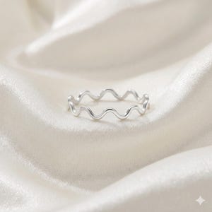 Könnte beinhalten: Ein silberfarbener Ring mit einem wellenförmigen Design. Der Ring besteht aus glänzendem Metall und hat ein durchgehendes Wellenmuster. Er wird auf einem weißen, satinartigen Stoff präsentiert.
