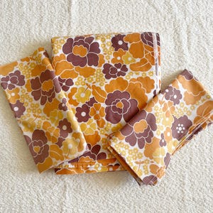 Pode incluir: Um conjunto de peças de tecido com um padrão floral repetitivo em tons de castanho escuro, amarelo mostarda e laranja claro sobre um fundo branco. O conjunto inclui uma peça retangular grande e duas peças retangulares dobradas menores.