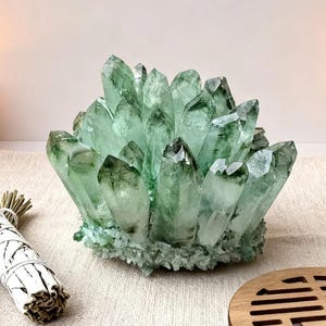 May include: A cluster of green crystals sits on a beige woven mat. The crystals have a natural, jagged appearance. A bundle of white sage and lavender sprigs are to the left of the crystals. A lit candle and a wooden coaster are also in the image.