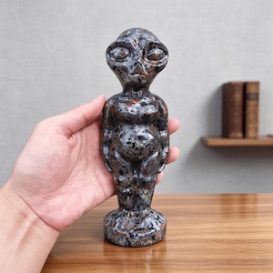 May include: A carved stone figurine depicting a humanoid alien with a large head, almond-shaped eyes, and a small nose. The sculpture features a mottled, polished surface with a colour palette of dark grey, black, and copper-brown mineral flecks.