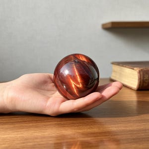 Puede incluir: Una piedra preciosa esférica y pulida de ojo de tigre rojo, con bandas de color caoba profundo y óxido, que presenta un brillo chatoyante y reflectante. La piedra redonda y lisa muestra vetas minerales naturales brillantes.