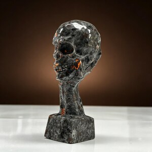 K&ouml;nnte beinhalten: Eine dunkelgraue Totenkopfskulptur mit orangefarbenen Akzenten. Der Sch&auml;del ist auf einem Hals und einem rechteckigen Sockel montiert. Die Skulptur hat eine polierte, reflektierende Oberfl&auml;che, vor braunem Hintergrund.