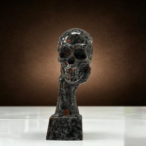 K&ouml;nnte beinhalten: Eine dunkelgraue Totenkopfskulptur mit orangefarbenen und braunen Akzenten. Der Sch&auml;del wird von einer Hand und einem Arm gehalten, die auf einem rechteckigen Sockel ruhen. Die Skulptur hat eine polierte, gl&auml;nzende Oberfl&auml;che.
