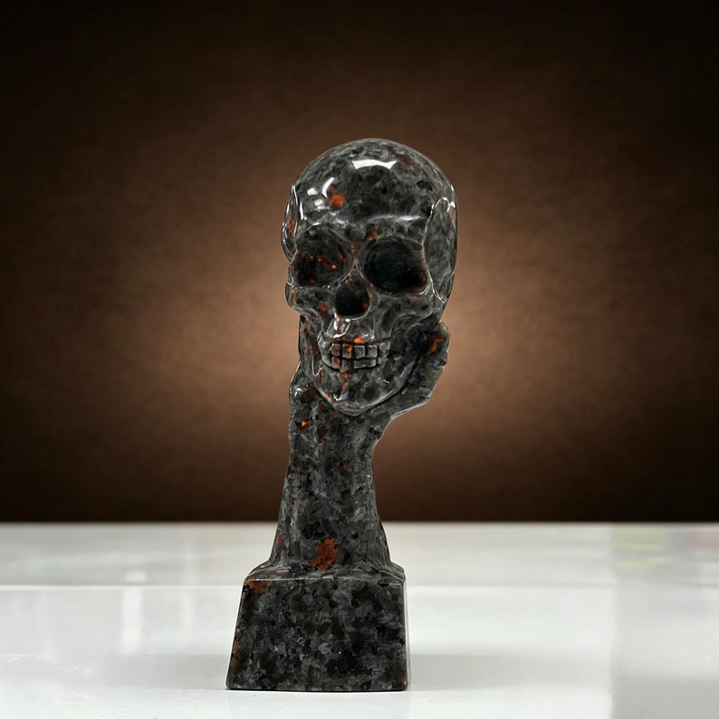 K&ouml;nnte beinhalten: Eine dunkelgraue Totenkopfskulptur mit orangefarbenen und braunen Akzenten. Der Sch&auml;del wird von einer Hand und einem Arm gehalten, die auf einem rechteckigen Sockel ruhen. Die Skulptur hat eine polierte, gl&auml;nzende Oberfl&auml;che.