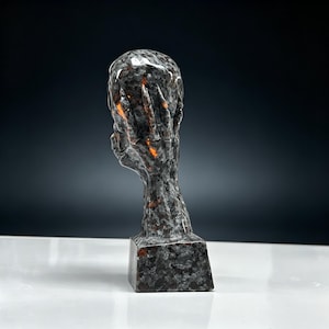 K&ouml;nnte beinhalten: Eine Skulptur eines Kopfes mit den H&auml;nden vor dem Gesicht. Die Skulptur besteht aus dunklem, grauem Stein mit orangefarbenen Akzenten. Sie steht auf einem quadratischen Sockel.